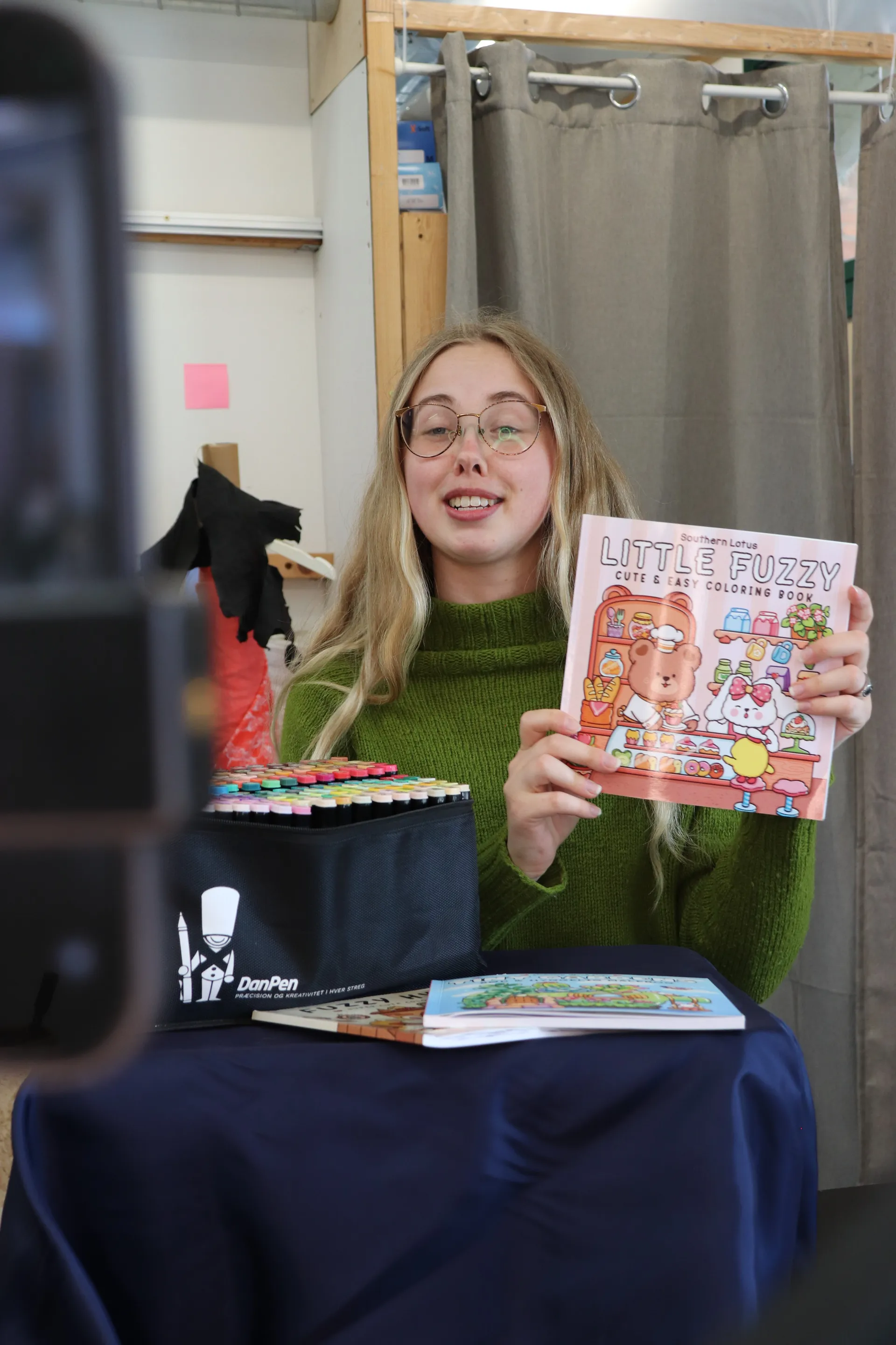 Vært holder en Little Fuzzy easy coloring book frem mod kameraet med tuscher på bordet