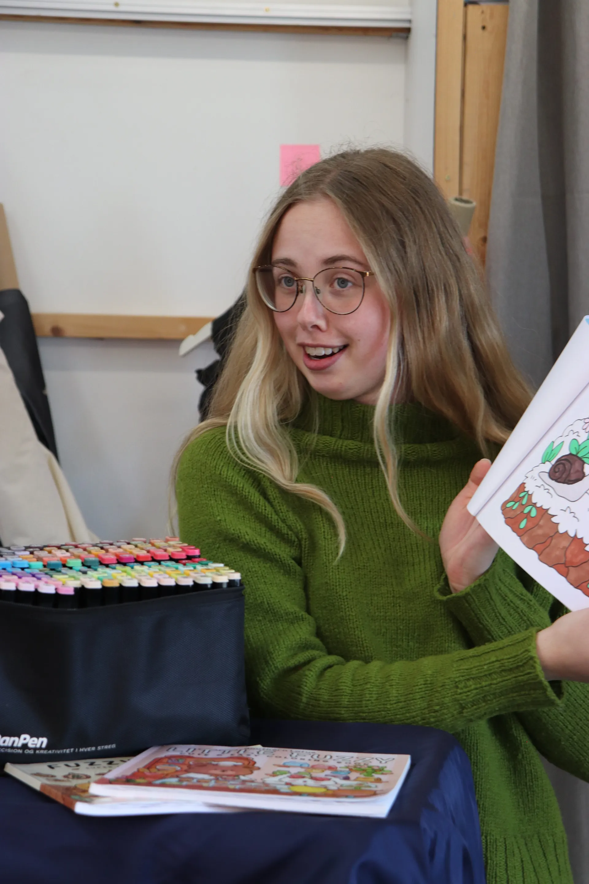 Vært holder en åben malebog frem og viser en grøntsagsillustration med tuscher på bordet