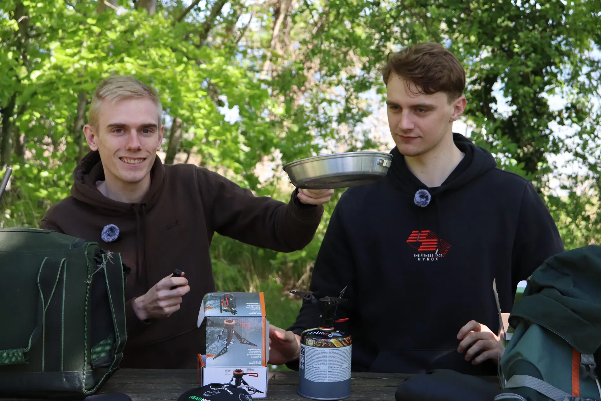 To værter ved et udendørs bord, den ene holder en titaniumpande op mens den anden betjener campingblusset