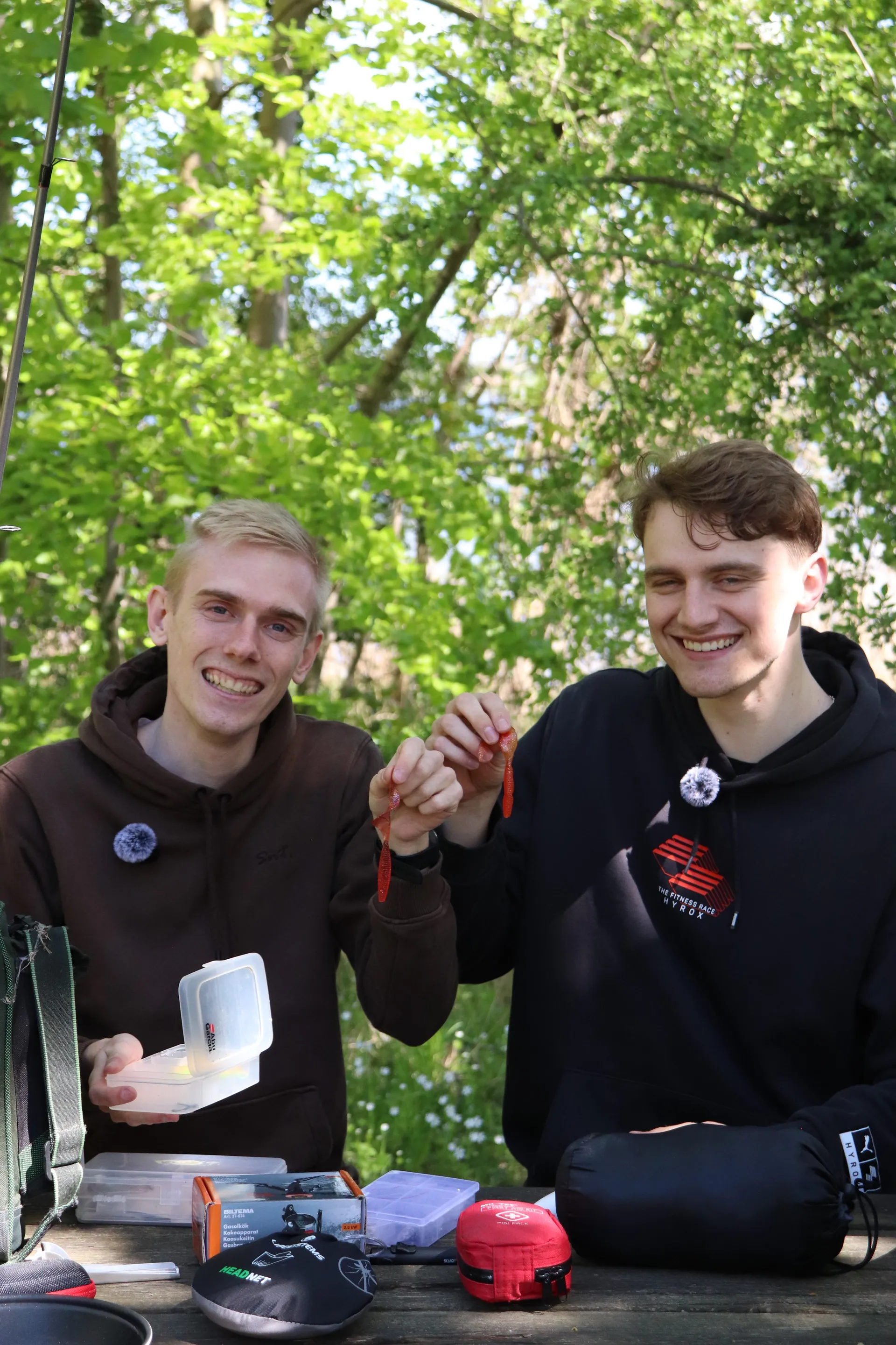 To værter viser friluftsgrej frem ved et campingbord i skoven under et live show